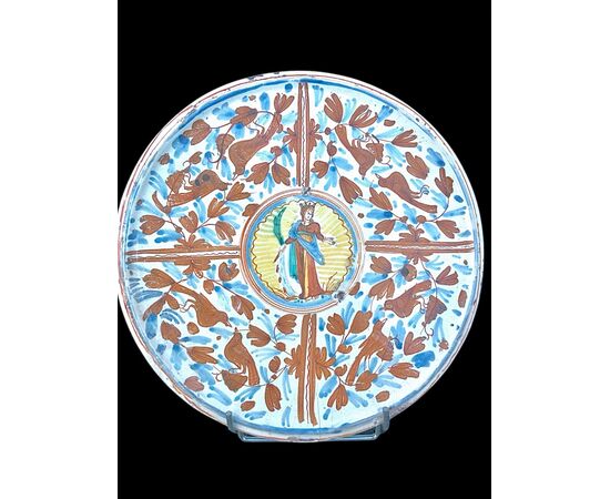 Alzata in maiolica a decoro calligrafico-naturalistico con al centro figura di Santa Caterina d’Alessandria che regge una palma, una spada e un libro.Deruta